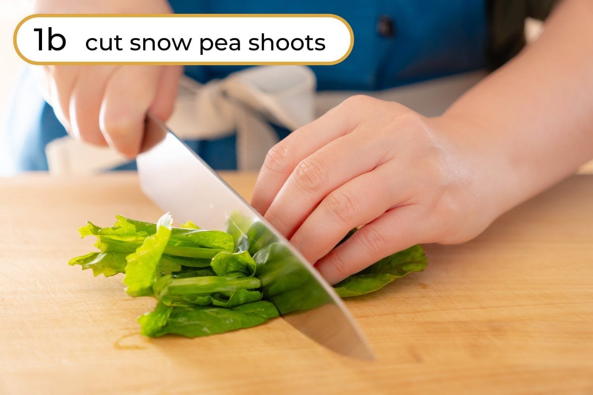 Cut pea shoots step 1a - cut pea shoots: Cindy cutting a small bunch of fresh pea shoots in half lengthwise on a wooden cutting board, with text "1b cut snow pea shoots" in the upper left corner.