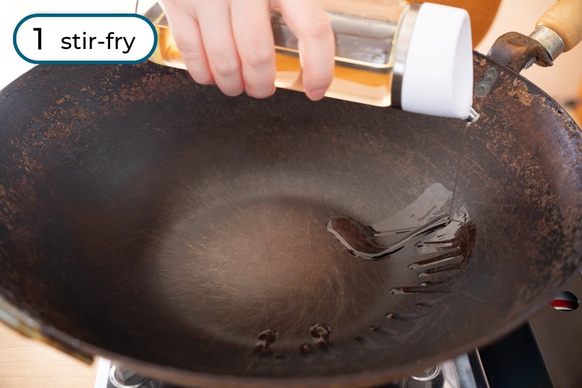 Stir-fried pea shoots step 1 - Cindy pours canola oil from a bottle into a large, heated, empty wok on a stove, preparing for stir-frying.
