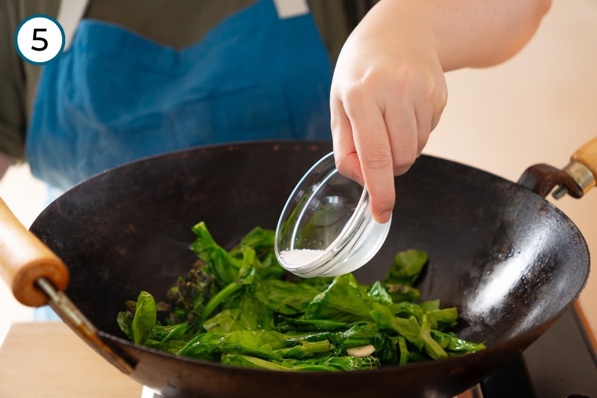 Stir-fried pea shoots step 5 - Cindy adding salt from a small glass bowl to pea shoots cooking in a wok.