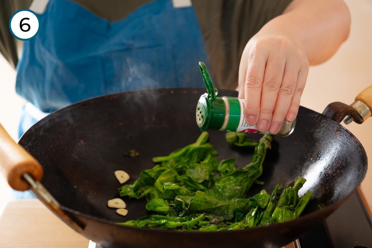 Stir-fried pea shoots step 6 - Cindy sprinkling ground white pepper onto the stir-fried pea shoots cooking in a wok.