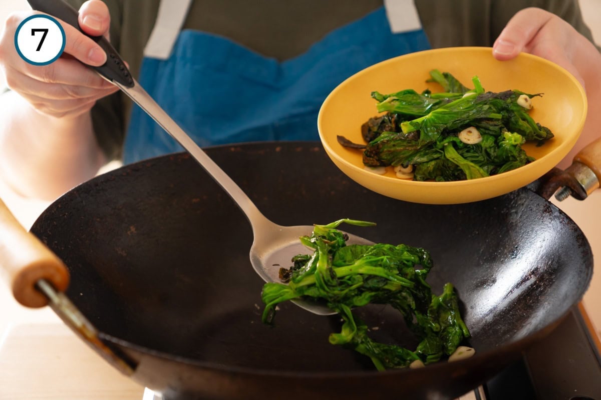 Stir-fried pea shoots step 7 - Cindy transferring the cooked vegetables from the wok into a yellow shallow serving bowl using a metal wok spatula.
