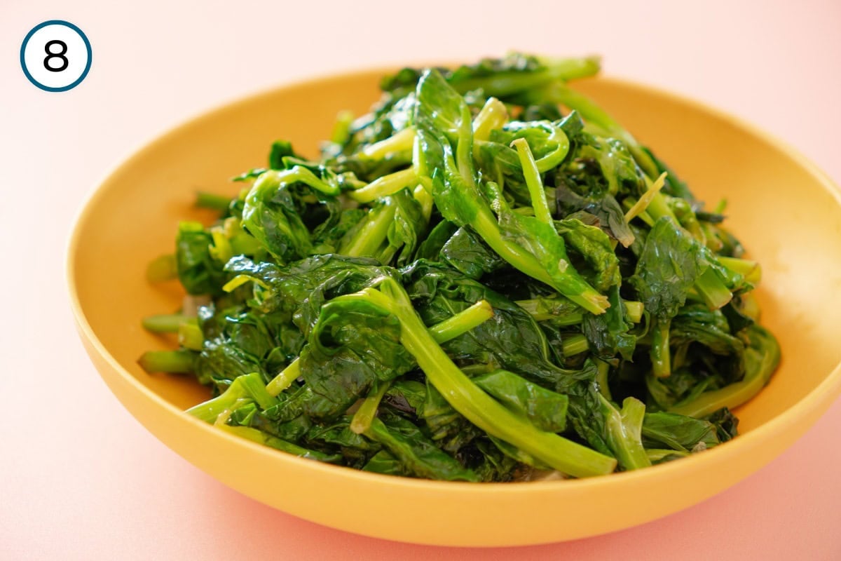 Stir-fried pea shoots step 8 - A shallow yellow serving bowl filled with cooked pea shoots sits on a pink surface.