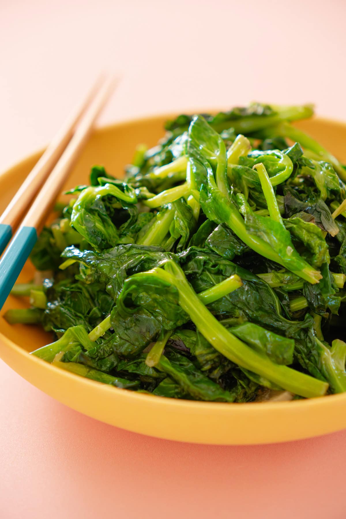 Stir-fried pea shoots in a shallow yellow serving bowl with a pair of chopsticks resting on the edge of the bowl, set against a pastel peach background.