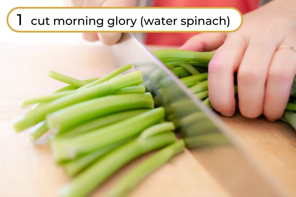Cut morning glory step 1 - Cindy cutting morning glory stems into shorter pieces with a chef's knife on a wooden board, with text "1 cut morning glory (water spinach)" in the upper left corner.