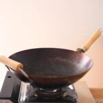 A black wok with wooden handles sits on a portable gas stove against a plain light-colored background.