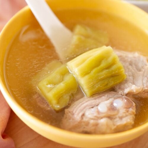 A bowl of clear bitter melon soup with chunks of green bitter melon, pieces of pork rib, and a white soup spoon.