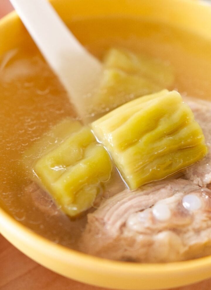 A bowl of clear bitter melon soup with chunks of green bitter melon, pieces of pork rib, and a white soup spoon.